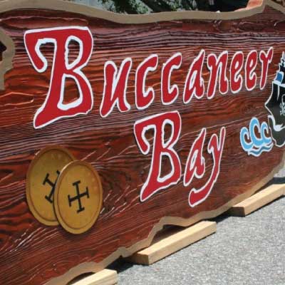Faux wood textured and stained solid wood sign with red and white outlined text with black pirate ship graphic. Text reads: Buccaneer Bay